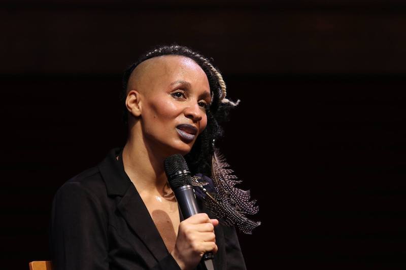 Adia Whitaker speaks into a microphone during the event's post-talk. She's wearing purple lipstick and has long braids and feathers that cascade down her right shoulder.